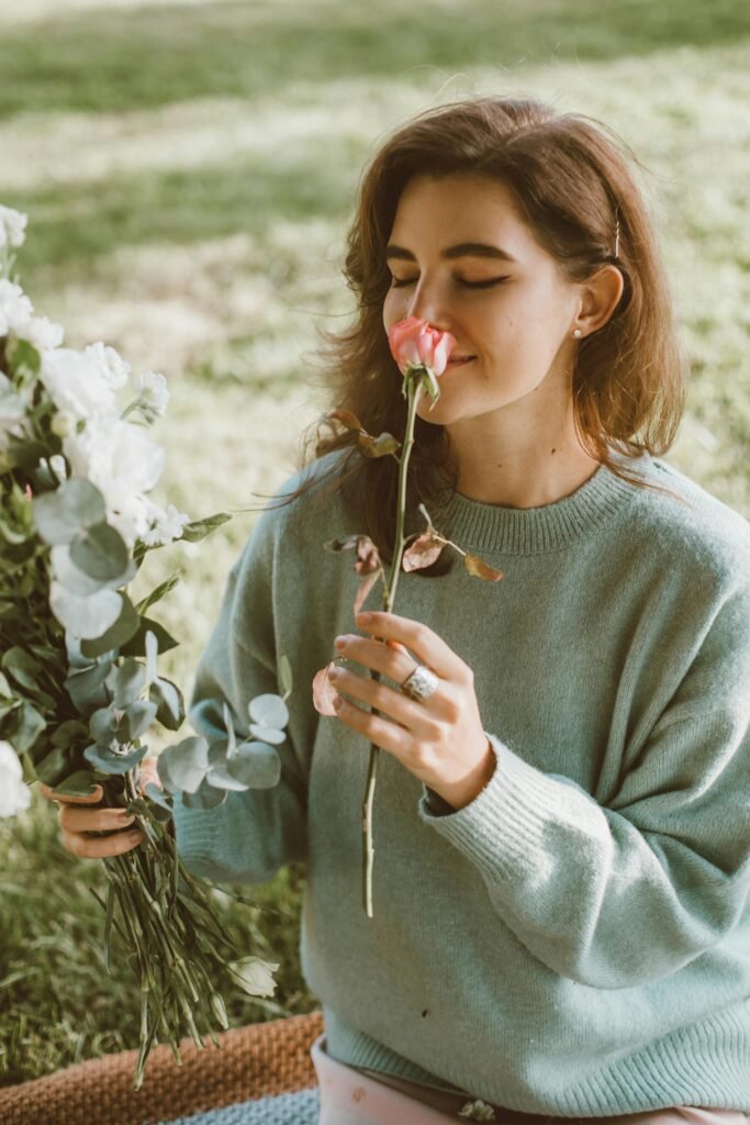 A woman in a sweater smells a pink rose while holding a flower bouquet outdoors.