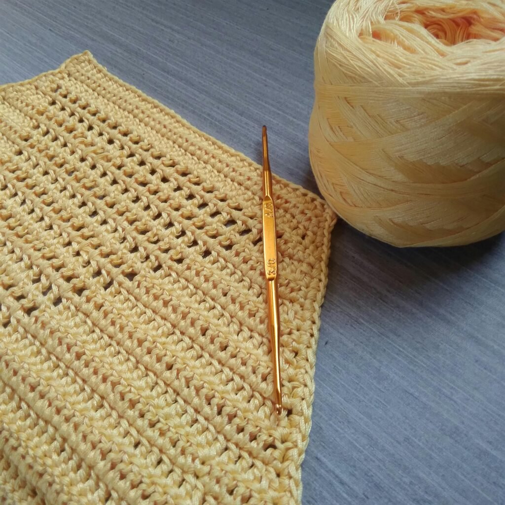 Close-up of a yellow crochet piece with a crochet hook and yarn ball on a blue surface.