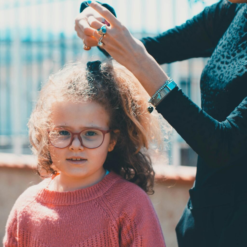 A young girl wearing glasses in a knitted sweater getting her hair styled outdoors on a sunny day.